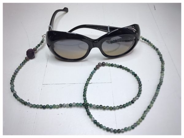 Łańcuszek do okularów z kamieni w kolorze ciemno zielonym z fioletowymi i szarymi dodatkami na lnianej torebce opakunkowej.
