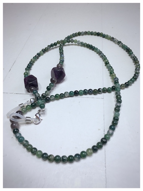 Łańcuszek do okularów z kamieni w kolorze ciemno zielonym z fioletowymi i szarymi dodatkami na lnianej torebce opakunkowej.