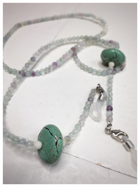 A chain for glasses made of celadon-violet glass stones with a celadon larger stone on a linen packaging bag.