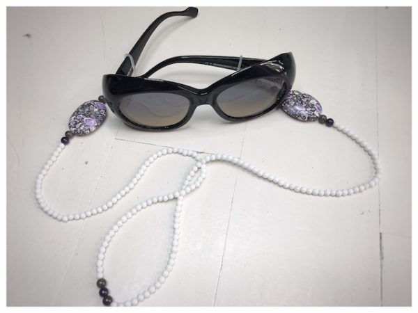 Łańcuszek do okularów z kamieni koloru białego i większymi biało-szaro-fioletowymi na lnianej torebce opakunkowej.
