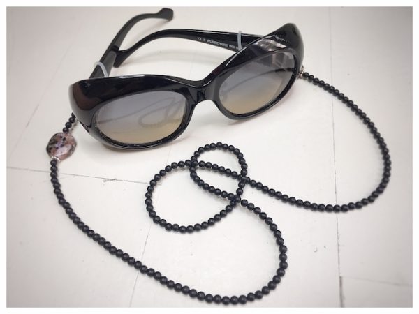 A chain for glasses made of black stones with one larger purple-black stone on a linen packaging bag.