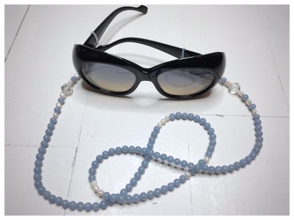 Łańcuszek do okularów dobro to obrazek jasnoniebieskiego łańcuszka z kamieni naturalnych z perłami i przezroczystymi kryształami na lnianym woreczku.